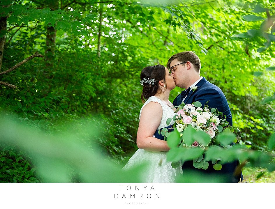 trevor and olivia kissing at their smithview pavillion wedding surrounded by lush green woods with leaves/branches out of focus in front of lens